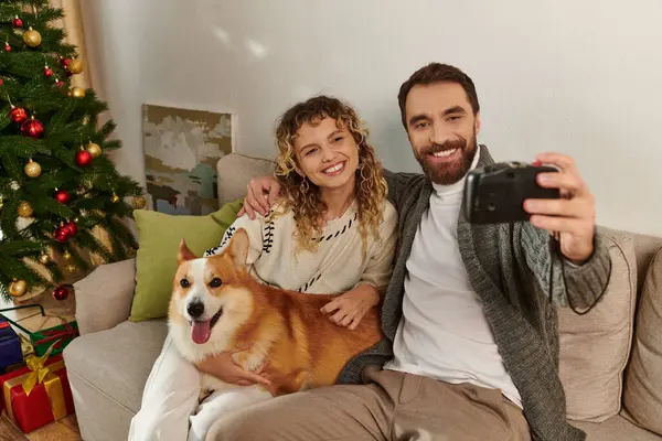 Happy couple smiling and taking photo on camera with corgi dog near decorated Christmas tree — Stock Photo