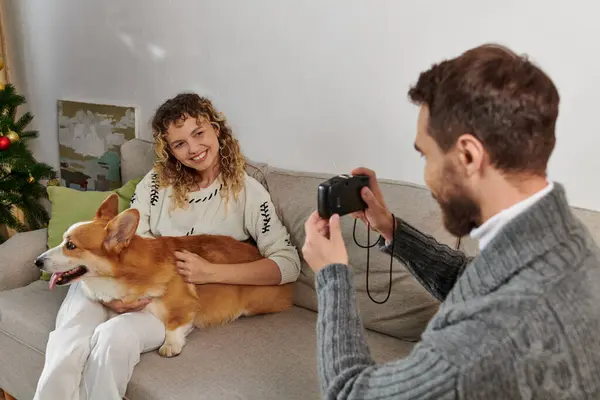 Happy man smiling and taking photo of wife with corgi dog on camera near Christmas tree — Stock Photo