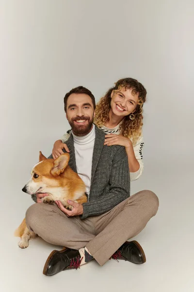 Cheerful man and woman in winter clothing smiling and playing with cute corgi on grey background — Stock Photo