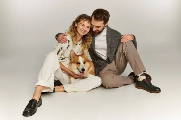 Cheerful man and woman in winter clothing smiling and playing with cute corgi on grey background — Stock Photo