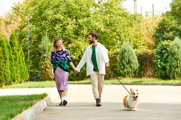 Feliz pareja en elegante atuendo cogido de la mano y caminando con lindo perro corgi alrededor de árboles verdes - foto de stock