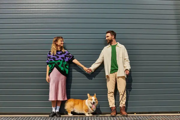 Feliz e elegante casal de mãos dadas e de pé com corgi cão perto de porta de garagem cinza — Fotografia de Stock