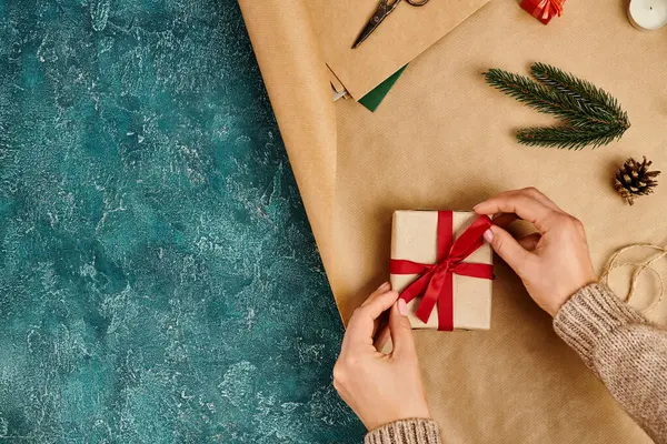 Cropped view of woman tying red ribbon bow on gift box near pine decor on blue textured backdrop — Stock Photo