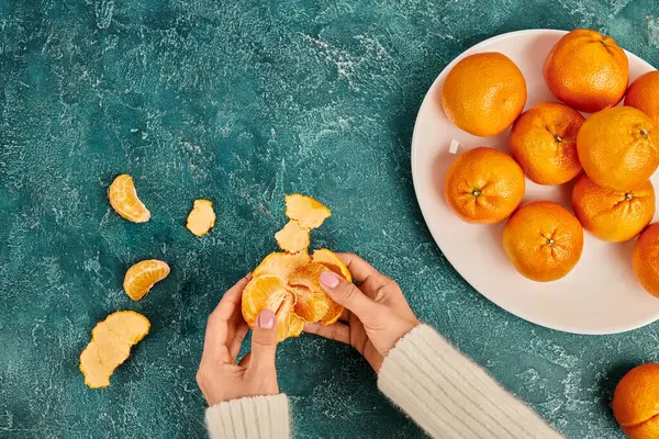 Cropped view of woman peeling fresh and sweet tangerine on blue textured backdrop, Christmas concept — Stock Photo