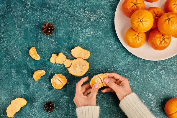 Vista cortada de mulher descascando tangerina suculenta perto de cones de pinho de fundo texturizado azul, Natal — Fotografia de Stock