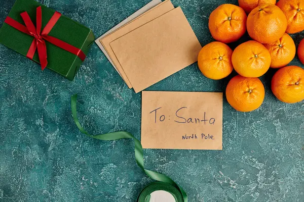 Лист Санті на північному полюсі біля мандаринів і подарункова коробка зі стрічкою на синьому текстурованому фоні — стокове фото