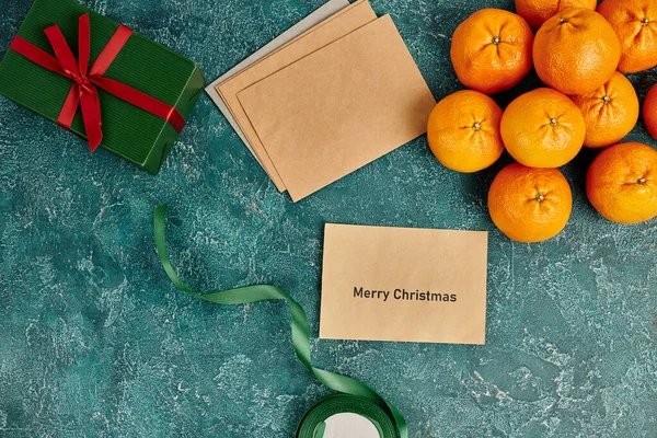 Sobre con letras de Feliz Navidad cerca de mandarinas y caja de regalo con cinta sobre fondo azul - foto de stock