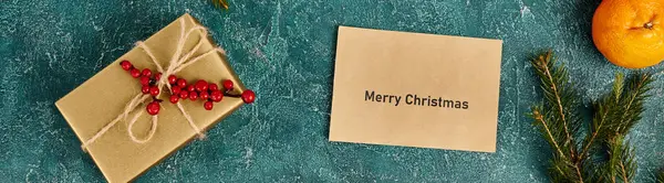 Merry Christmas envelope near mandarin and gift box with holly berries on blue texture, banner — Stock Photo
