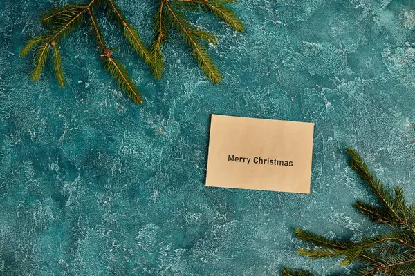 Поздравительный конверт с рождественской надписью рядом с сосновыми ветвями на синем текстурированном фоне — стоковое фото
