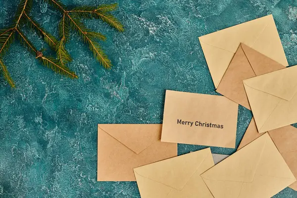 Craft paper envelopes near green pine branches on blue textured backdrop, Merry Christmas lettering — Stock Photo