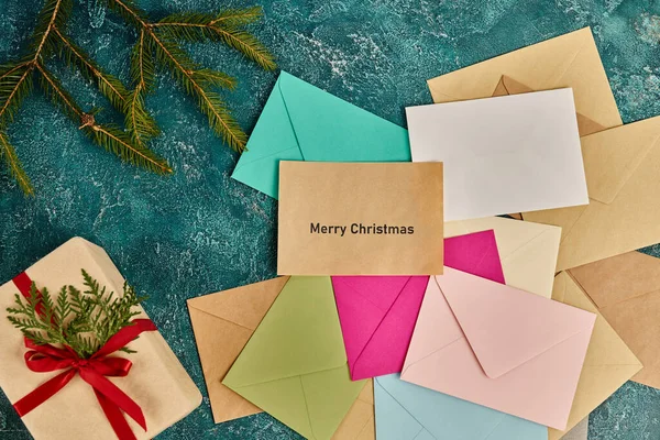 Présent près de branches de pin et enveloppes colorées sur fond bleu rustique, Joyeux Noël lettrage — Photo de stock