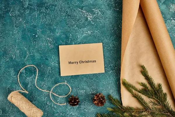 Busta con scritte Buon Natale vicino alla carta artigianale e spago su superficie blu con decorazioni in pino — Foto stock