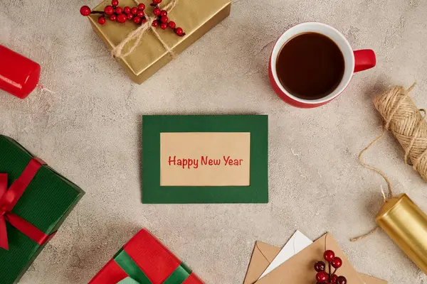 Heureuse nouvelle carte de vœux de l'année près de boîtes-cadeaux décorées et tasse de chocolat chaud sur surface texturée — Photo de stock