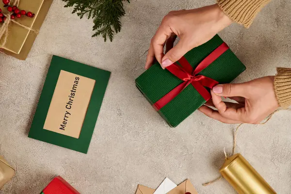 Vista recortada de la mujer atando lazo rojo en caja de regalo cerca de la tarjeta de felicitación de Feliz Navidad y decoración festiva - foto de stock