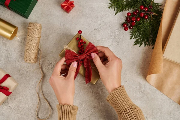 Vista recortada de la mujer decorando caja de regalo con lazo rojo cerca de suministros de artesanía, telón de fondo de Navidad - foto de stock