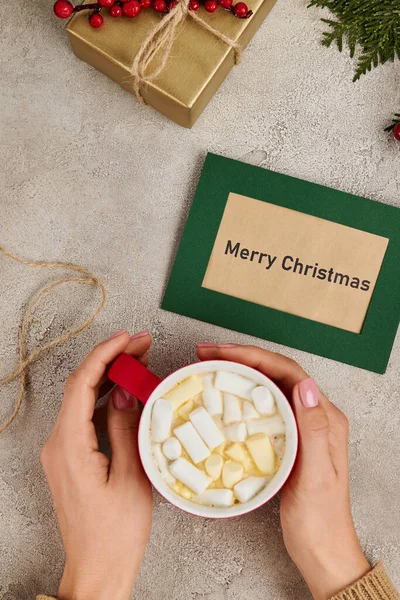 Partial view of woman holding hot chocolate with marshmallow near Merry Christmas greeting card — Stock Photo