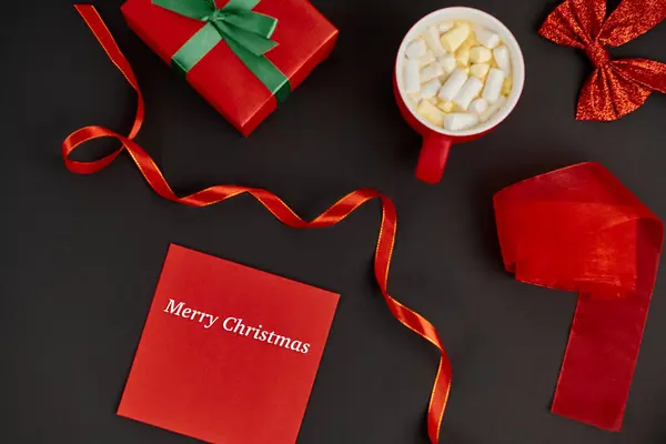 Красный конверт с рождественскими надписями возле коробки и горячий шоколад с зефиром на черном — стоковое фото