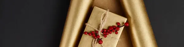 Shiny gift box with red holly berries on golden paper and black backdrop, horizontal banner — Stock Photo