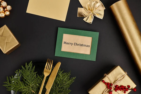 Greeting card with Merry Christmas lettering near golden cutlery and shiny festive decor on black — Stock Photo