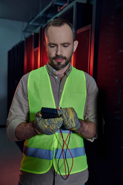 Attractive bearded man in safety vest and gloves looking at detector in his hands, data center — Stock Photo