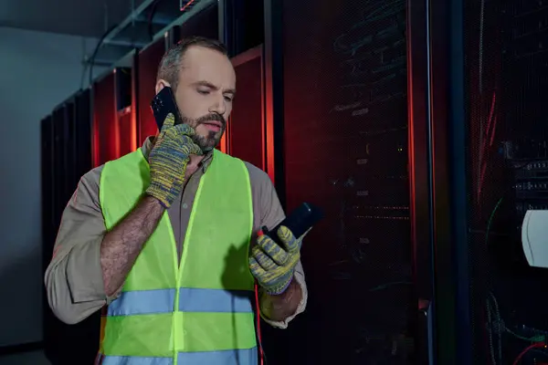 Handsome technician with beard in safety vest and gloves holding detector and talking by phone — Stock Photo