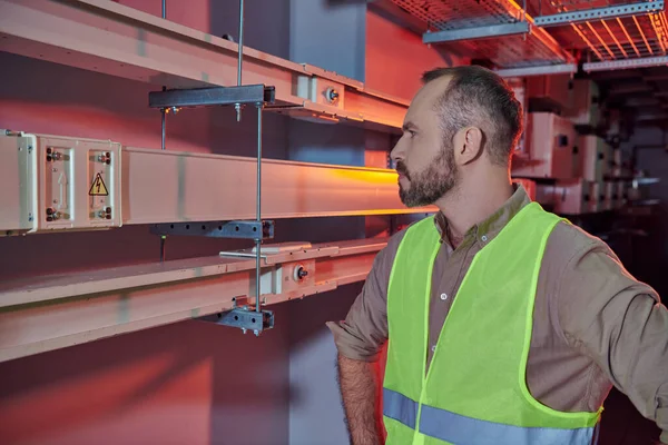 Handsome bearded specialist in safety vest with arms akimbo working attentively with data center — Stock Photo