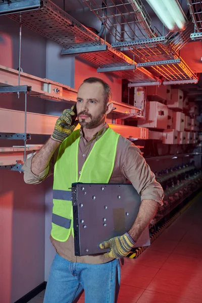 Concentrated technician in safety vest and gloves talking by phone and looking away during work — Stock Photo