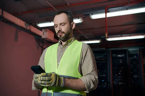Bello barbuto tecnico in giubbotto di sicurezza e guanti guardando il suo smartphone durante il lavoro — Foto stock