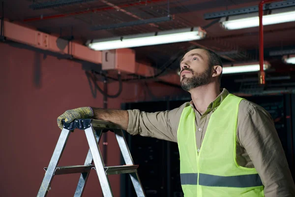 Apuesto técnico pensativo en chaleco de seguridad de pie junto a la escalera de paso y mirando hacia arriba, centro de datos - foto de stock