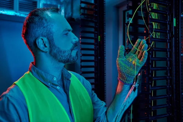 Gut aussehender, nachdenklicher Techniker in Sicherheitskleidung, der im Rechenzentrum mit Kabeln und Drähten arbeitet — Stockfoto