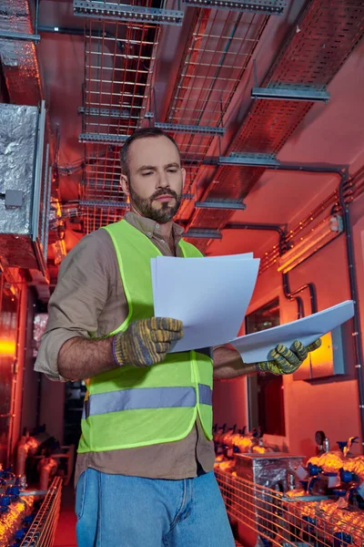 Handsome focused specialist in safety gloves and vest looking at his paperwork during inspection — Stock Photo