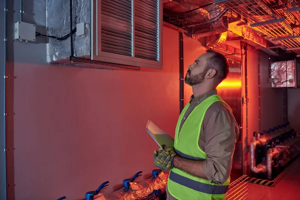 Attractive concentrated technician in safety gloves and vest holding papers and looking up, data — Stock Photo