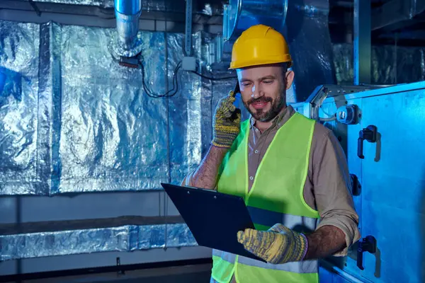 Jolly hardworking technician in safety helmet and clothes looking at clipboard and talking by phone — Stock Photo