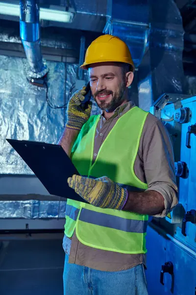 Joyous professional in safety vest and helmet looking at clipboard and talking by phone, data center — Stock Photo