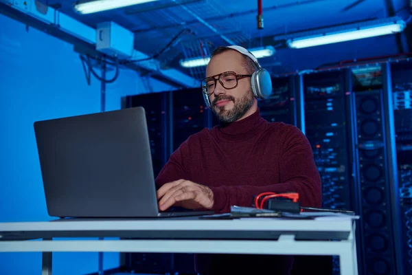 Allegro specialista attraente in dolcevita con cuffie che lavorano sul suo computer portatile, data center — Foto stock