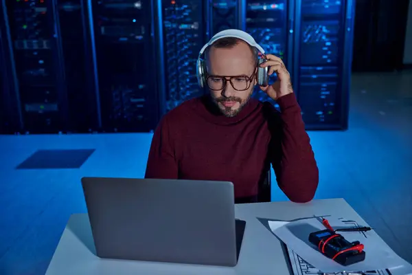 Attraente specialista focalizzato con barba e cuffie che lavorano duramente sul suo computer portatile, data center — Foto stock