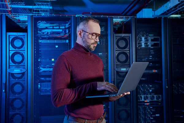Attractive bearded specialist in turtleneck working attentively at his laptop, data center — Stock Photo