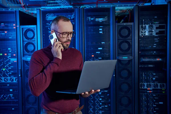 Pensive data center specialist in turtleneck with glasses talking by phone and looking at his laptop — Stock Photo
