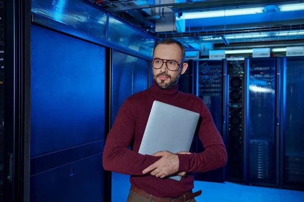 Good looking focused specialist with beard and glasses holding laptop and looking away at work — Stock Photo