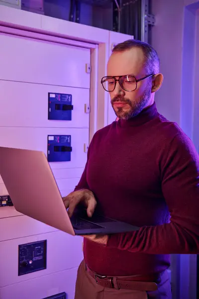 Attractive focused specialist in turtleneck with beard and glasses looking at his laptop attentively — Stock Photo