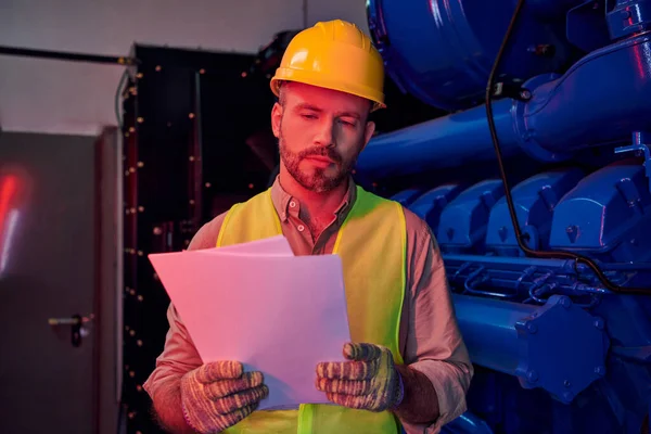 Concentrated devoted technician in safety vest and helmet looking at his paperwork, data center — Stock Photo
