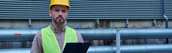 Pensive technician in helmet with clipboard and looking at camera while outside, data center, banner — Stock Photo