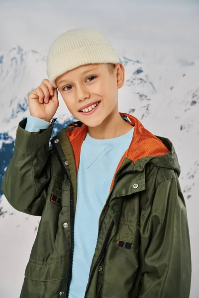Vertical shot of cheerful little boy in warm attire with hand on beanie hat smiling at camera — Stock Photo