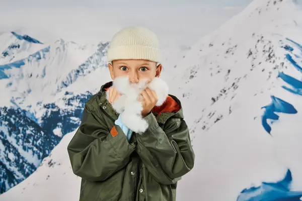 Cute little boy in beanie hat posing with snow in hands near face, looking at camera, fashion — Stock Photo