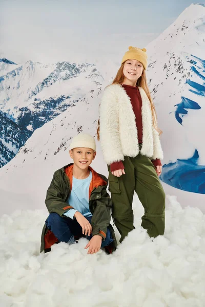 Vertical shot of little boy sitting on snow near cute standing girl, stylish outfit, winter fashion — Stock Photo