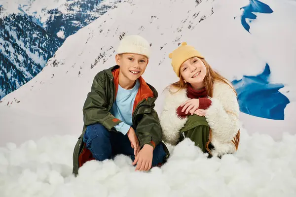 Niño y niña alegre sentado en la nieve en trajes cálidos y elegantes y sonriendo a la cámara, la moda - foto de stock