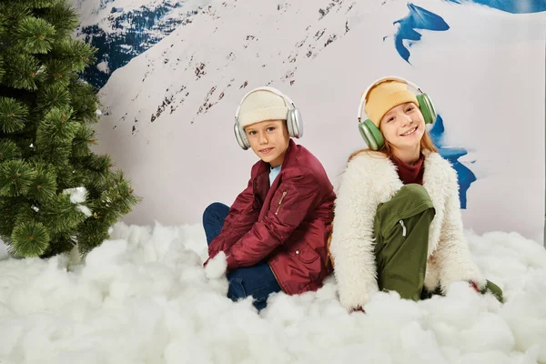 Niedlichen Jungen und Mädchen in stilvollen Winter-Outfits mit Headsets sitzen auf Schnee mit Bergkulisse — Stockfoto