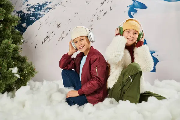 Jolly preteen amici in eleganti giacche seduti sulla neve toccando cuffie e sorridendo alla fotocamera — Foto stock