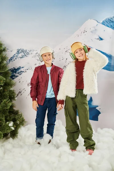 Счастливые дети в модных одеждах с наушниками, стоящими на снегу и весело улыбающимися, мода — стоковое фото