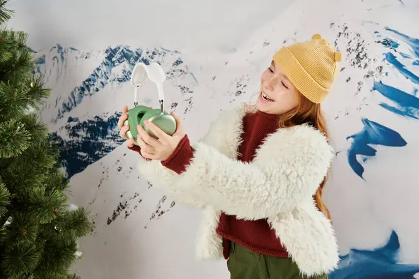 Feliz niña preadolescente en invierno elegante traje al lado de abeto mirando sus auriculares, concepto de moda - foto de stock